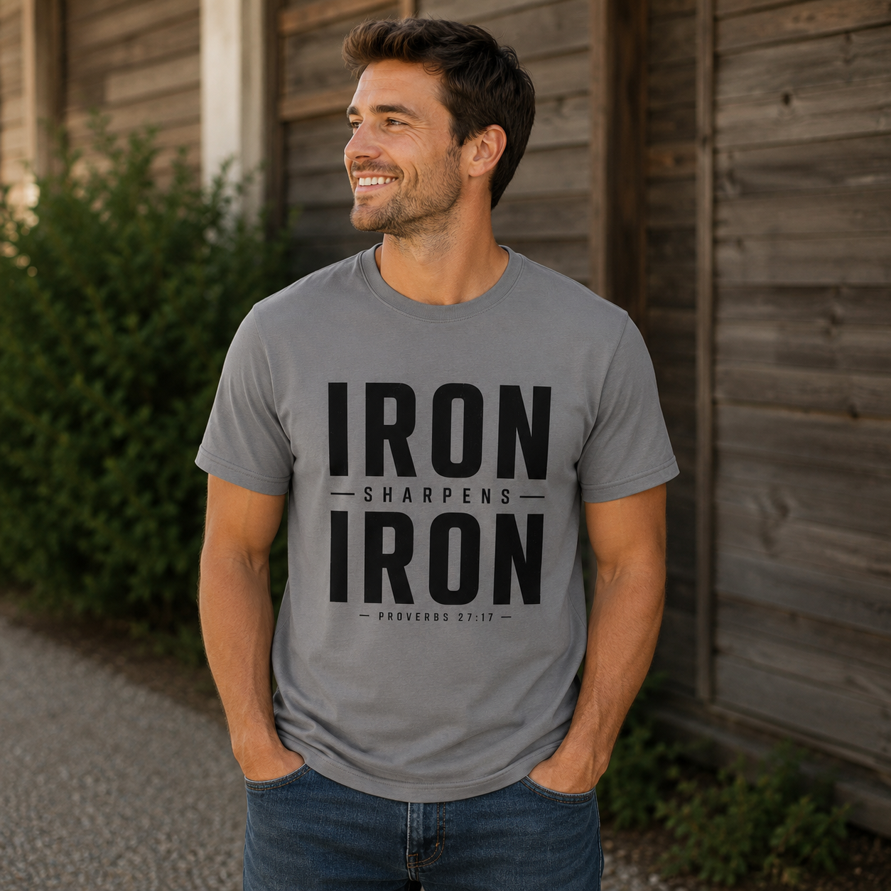 A man wearing a heather gray crewneck t-shirt with a black graphic print that reads 'IRON SHARPENS IRON' and 'PROVERBS 3:7-9' on the front, standing outdoors in front of a wooden wall.