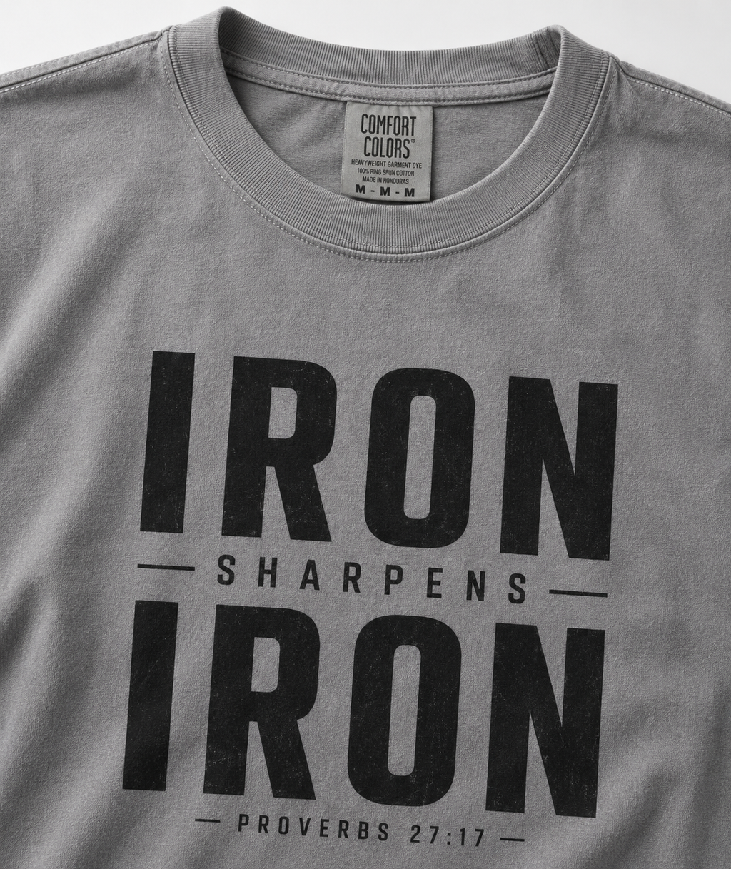 A close-up of a gray Comfort Colors t-shirt featuring a bold black graphic print that reads 'IRON SHARPENS IRON' with 'PROVERBS 27:17' underneath.